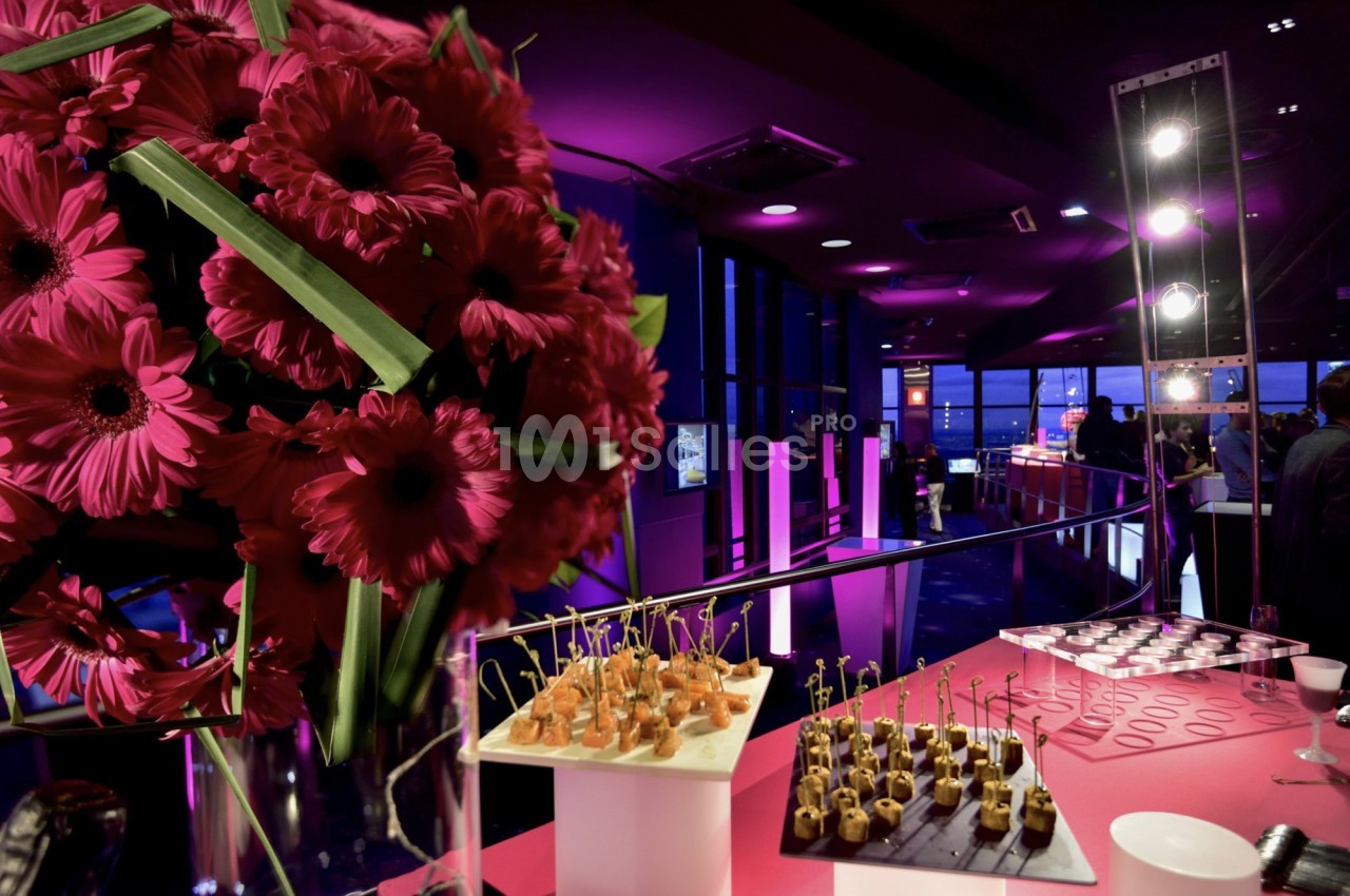 Bouquet de fleurs roses au premier plan avec un buffet de bouchées apéritives dans une salle éclairée en lumière violette.