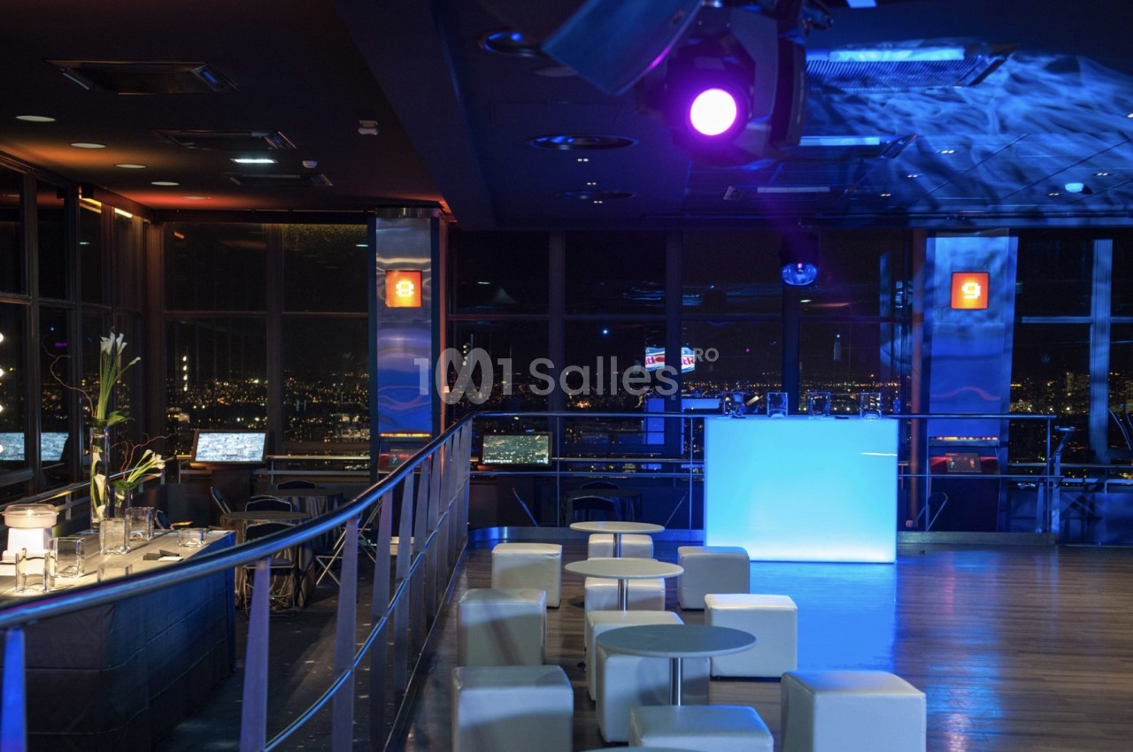 Salle moderne avec éclairage tamisé, tables basses blanches, bar et vue nocturne sur une ville illuminée.