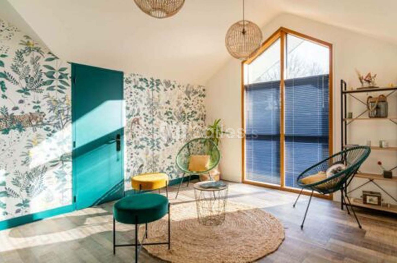 Salon lumineux avec mur végétal, grandes fenêtres, fauteuils modernes, tabourets colorés et étagère décorative.