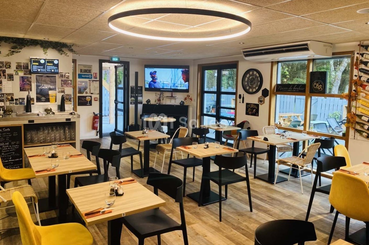 Salle de restaurant moderne avec tables en bois, chaises colorées et grande baie vitrée laissant entrer la lumière naturelle.