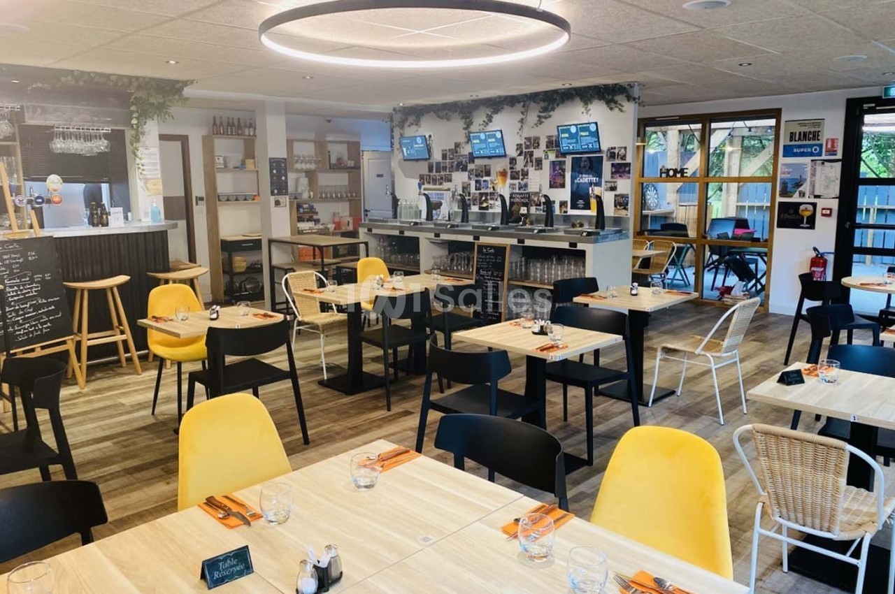 Salle de restaurant lumineuse avec tables en bois clair, chaises noires et jaunes, et un bar en arrière-plan.