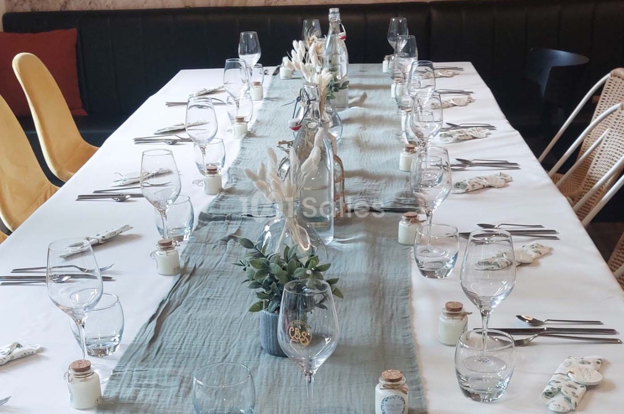 Table dressée avec une nappe blanche, chemin de table bleu, verres, couverts et décorations végétales.