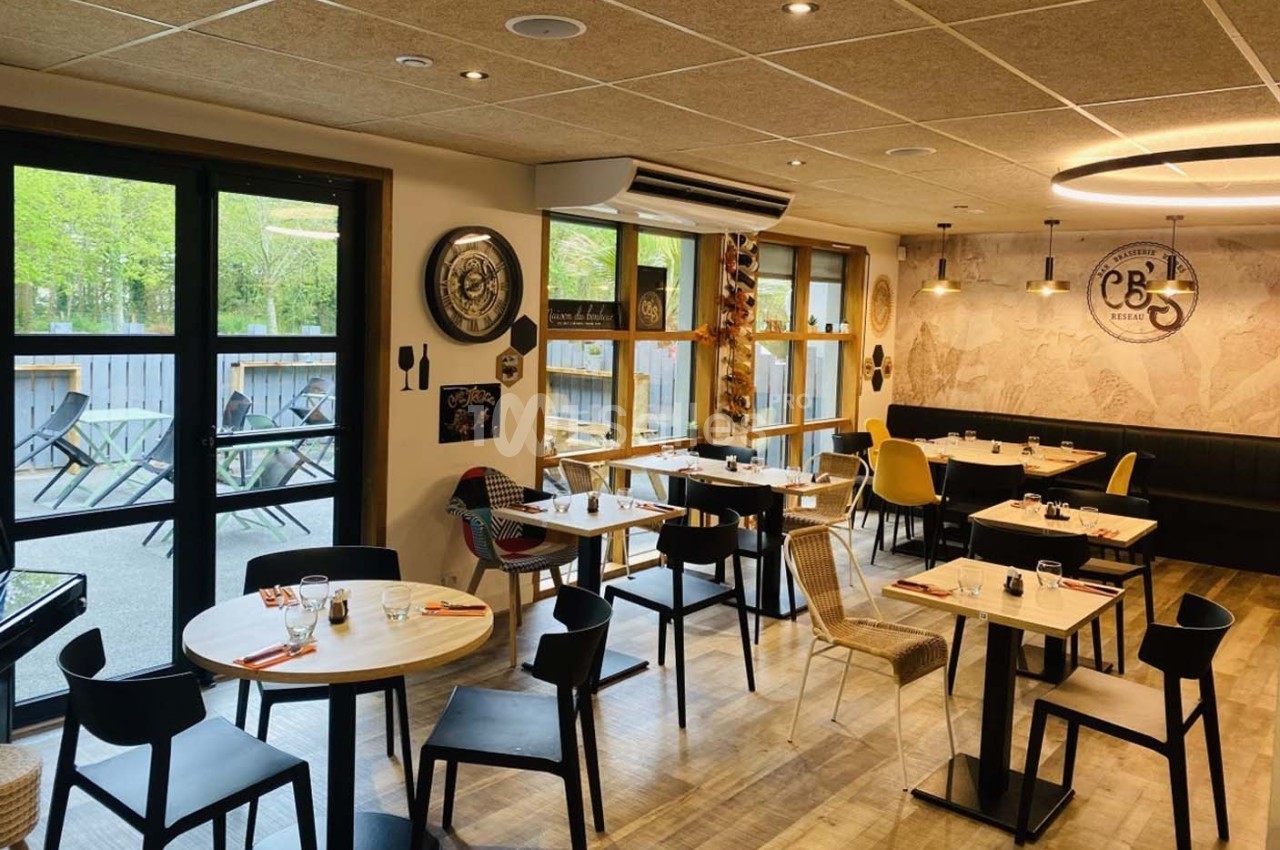 Salle de restaurant lumineuse avec tables dressées, chaises variées et vue sur une terrasse extérieure arborée.