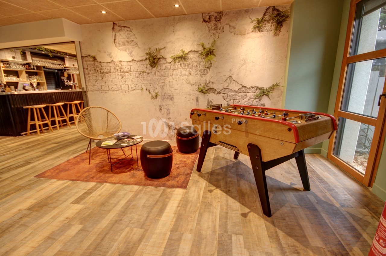Espace détente avec baby-foot, fauteuil en rotin, poufs et mur décoratif végétalisé dans un intérieur chaleureux.