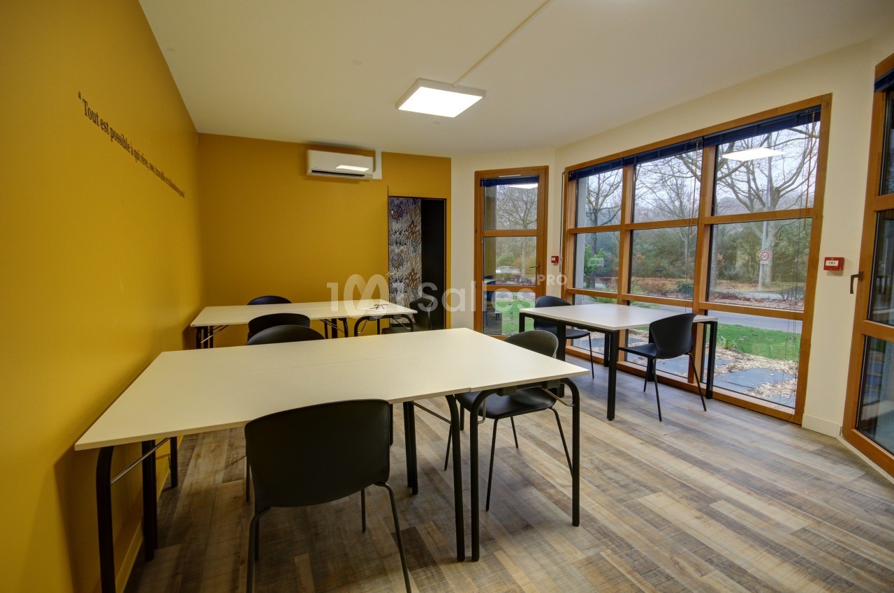 Salle lumineuse avec murs jaunes, tables et chaises noires, grandes fenêtres donnant sur un espace extérieur arboré.