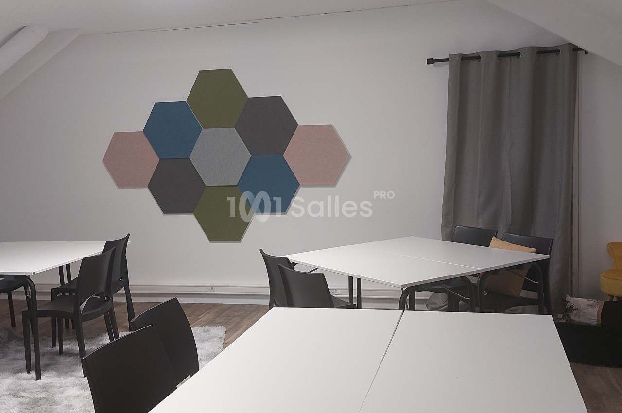 Salle lumineuse avec tables blanches, chaises noires, décoration murale hexagonale et coin détente avec fauteuil jaune.