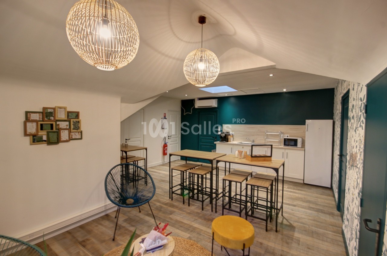Salle commune lumineuse avec cuisine équipée, tables hautes, chaises et décoration murale moderne.