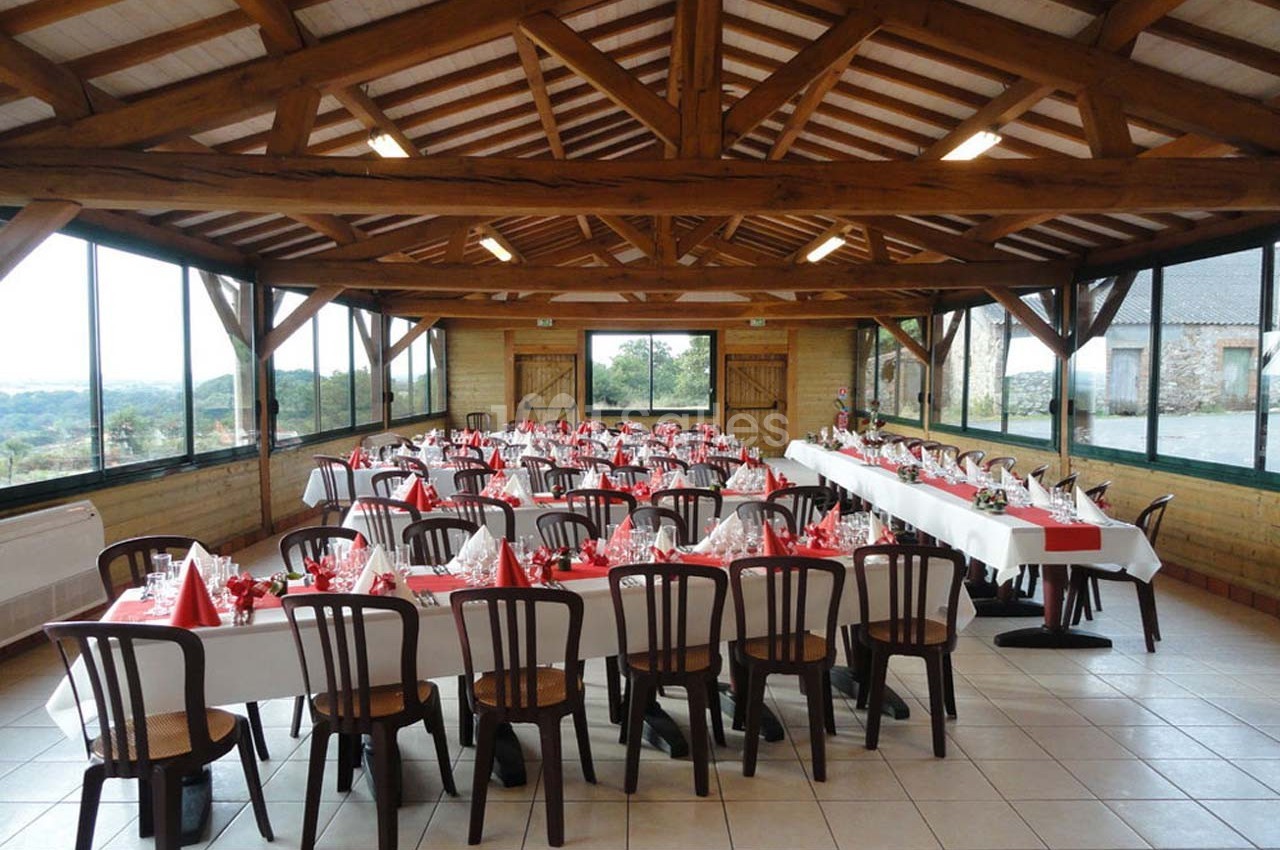 Salle de réception avec tables dressées, nappes blanches et décorations rouges, entourée de grandes baies vitrées.