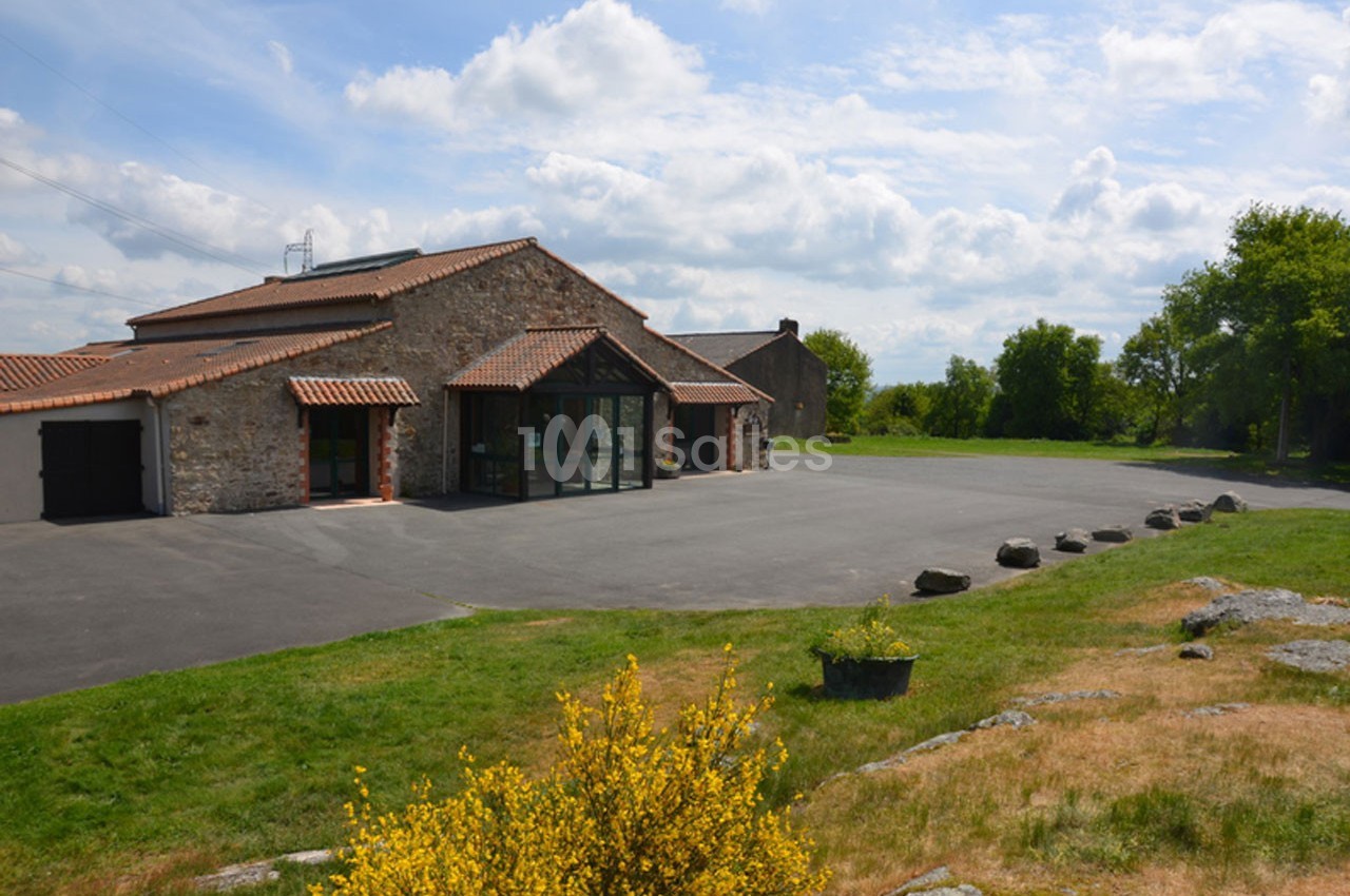 Bâtiment en pierre avec toit en tuiles, entouré d'une grande cour asphaltée et d'espaces verts parsemés de buissons.