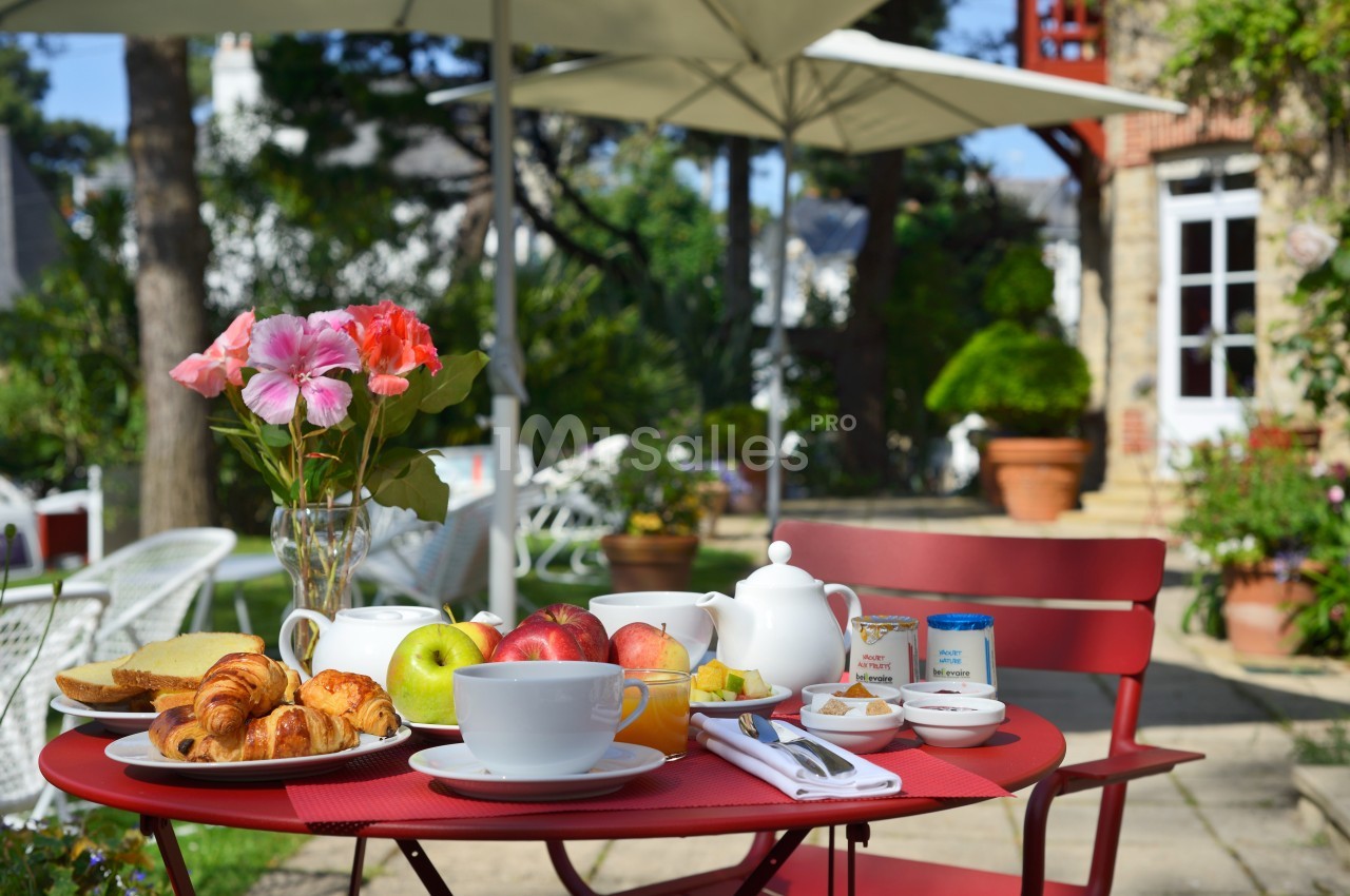 Petit-déjeuner servi en extérieur sur une table rouge avec viennoiseries, fruits, théière et fleurs en arrière-plan.