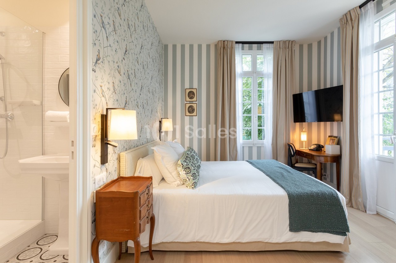 Chambre lumineuse avec lit double, bureau, télévision, salle de bain attenante et grandes fenêtres donnant sur un jardin.