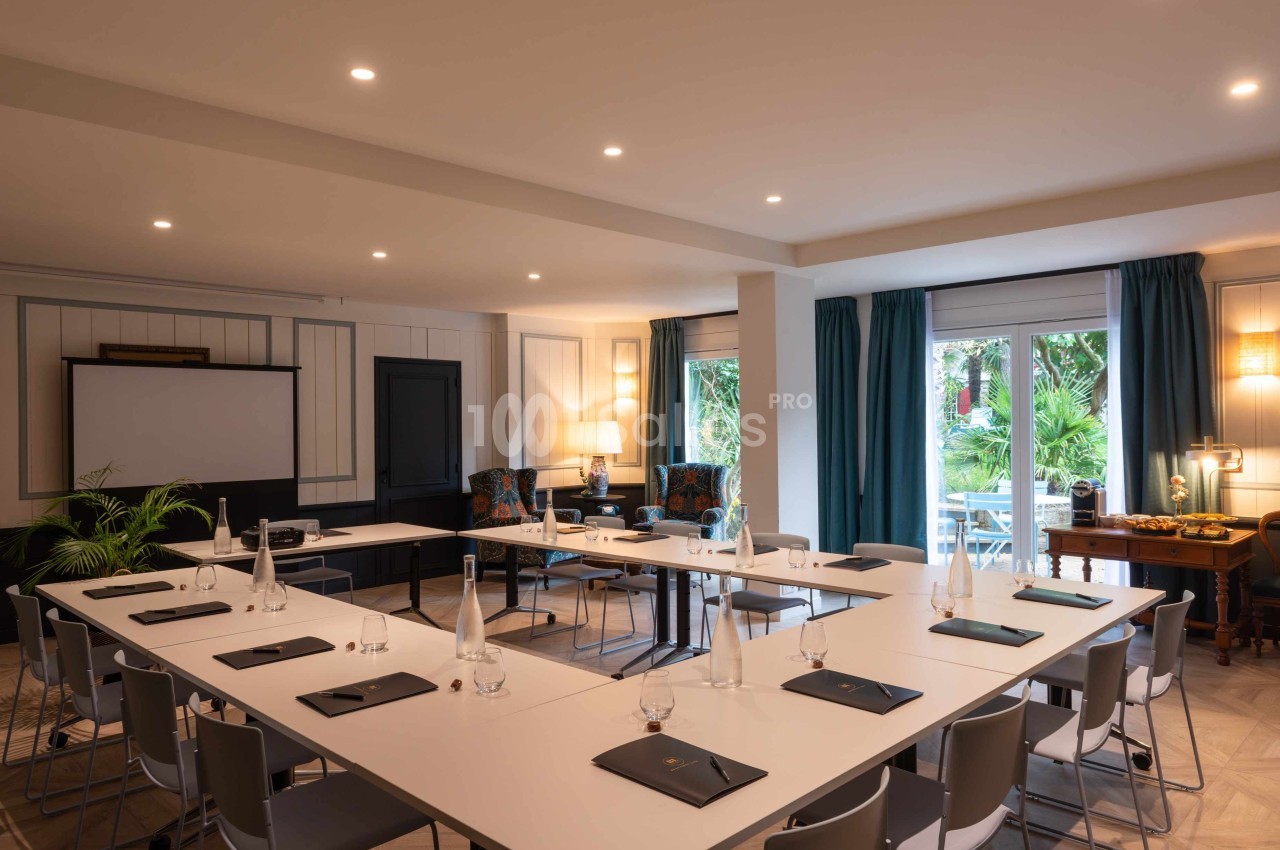 Salle de réunion lumineuse avec tables disposées en U, chaises grises, écran de projection et vue sur un jardin.