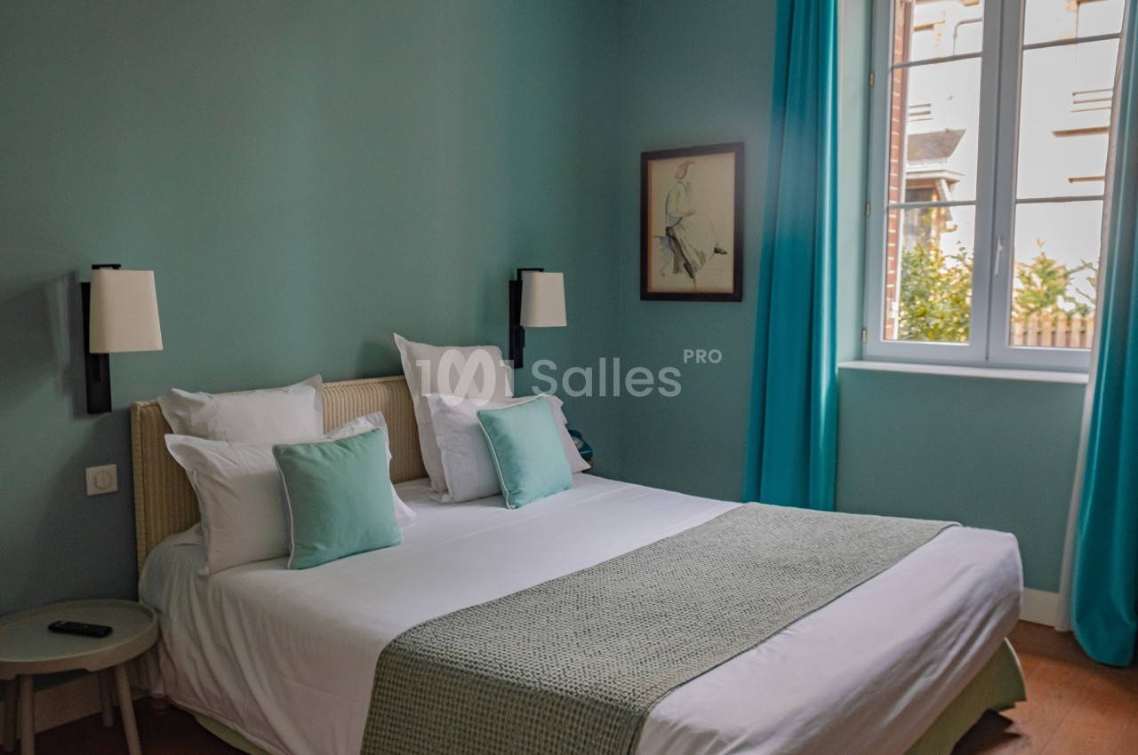 Chambre lumineuse avec lit double, coussins assortis, murs bleu clair, rideaux turquoise et fenêtre donnant sur l'extérieur.