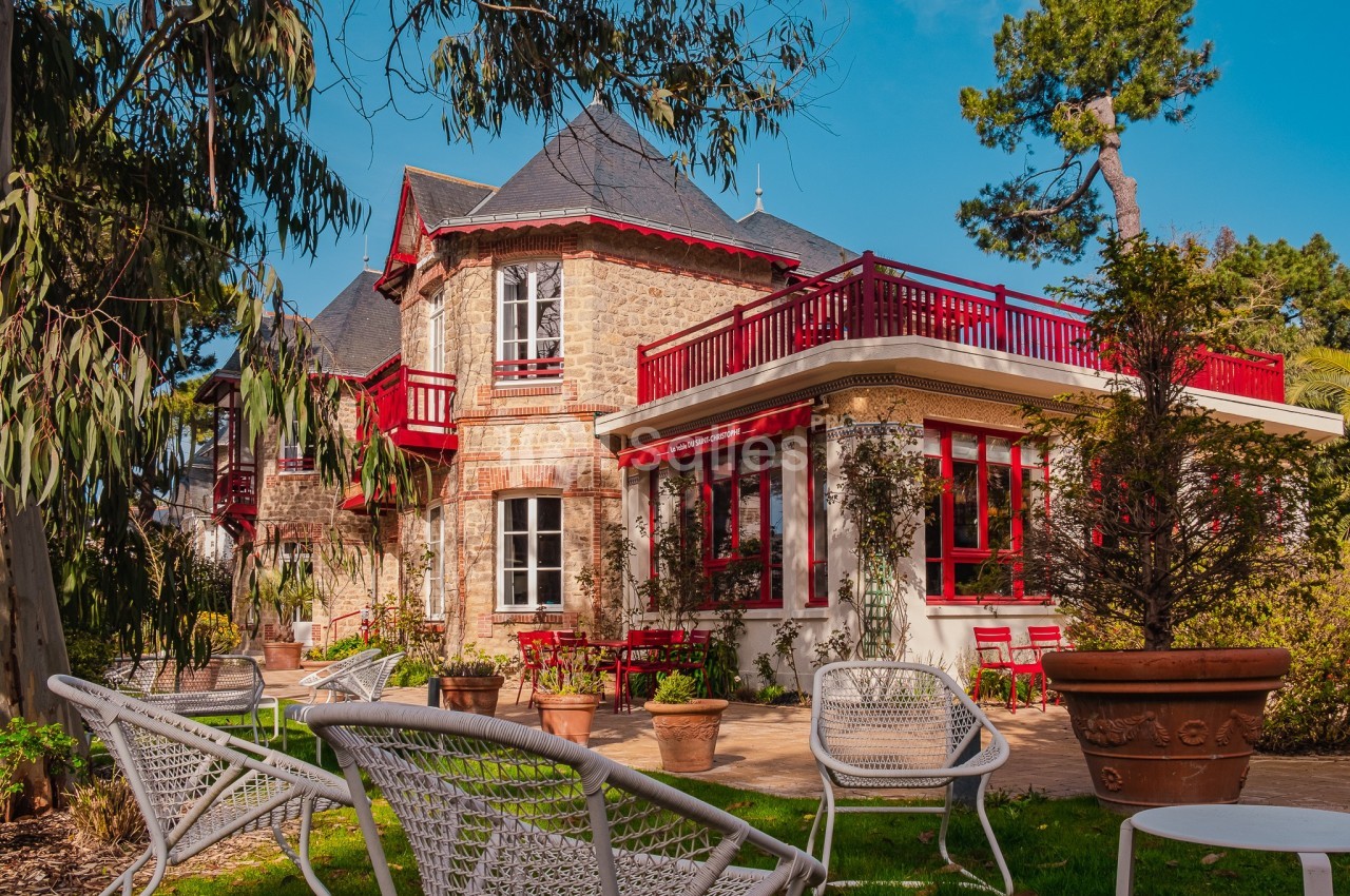 Maison en pierre avec balcons et volets rouges, entourée de végétation et de mobilier de jardin en métal blanc.