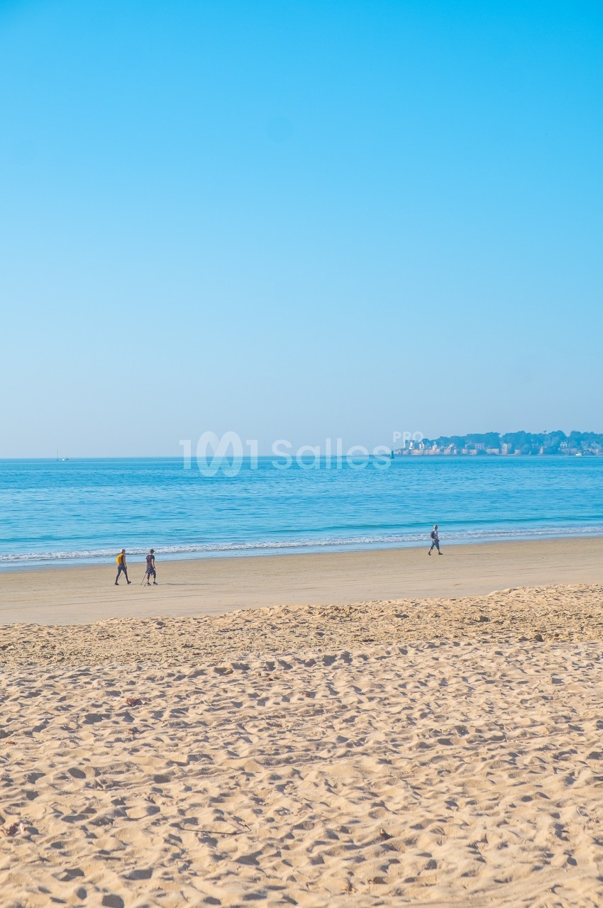 Trois personnes marchant sur une plage de sable fin avec une mer calme et un ciel bleu en arrière-plan.