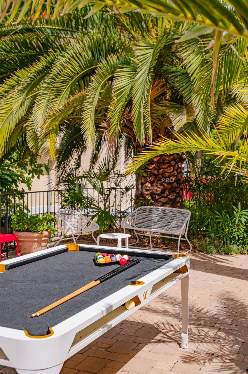Table de billard extérieure blanche avec accessoires, placée sur une terrasse pavée entourée de palmiers et de mobilier de…