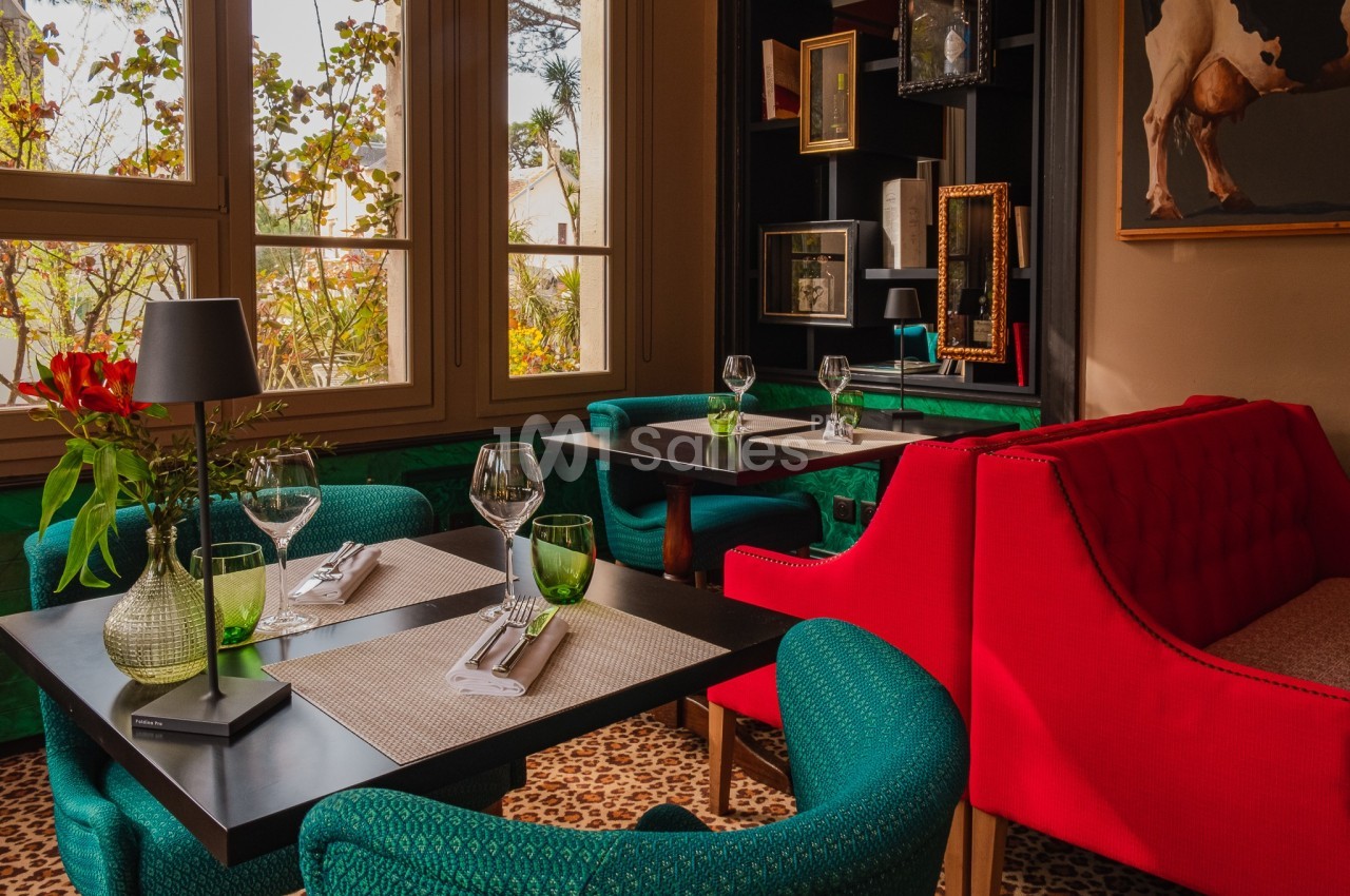 Salle de restaurant lumineuse avec tables dressées, fauteuils rouges et verts, et vue sur un jardin à travers de grandes…