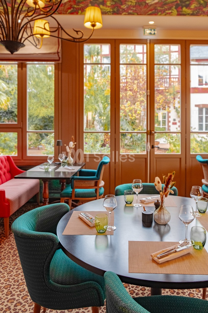 Salle de restaurant lumineuse avec tables dressées, chaises vertes et grandes baies vitrées donnant sur un jardin.