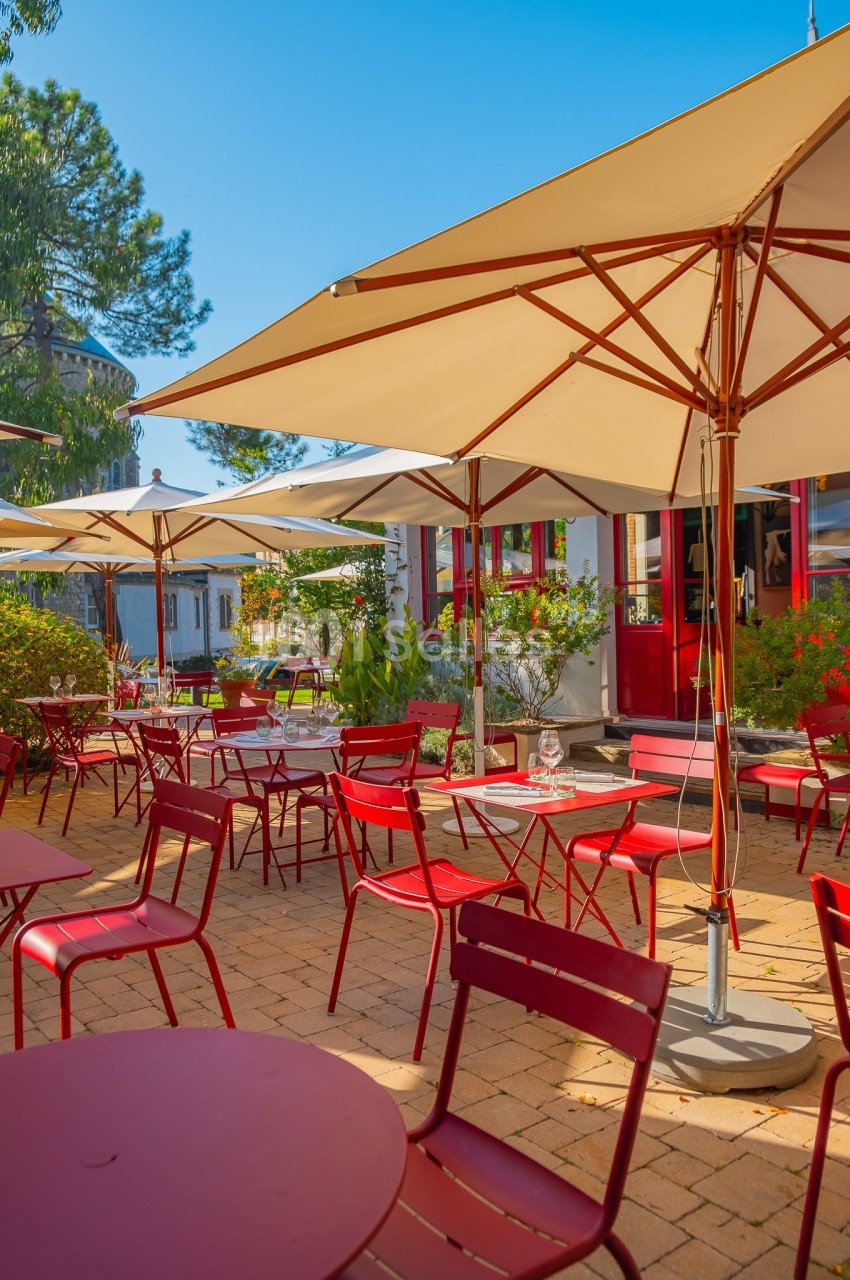 Terrasse ensoleillée avec tables et chaises rouges, entourée de parasols et de végétation, devant un bâtiment.