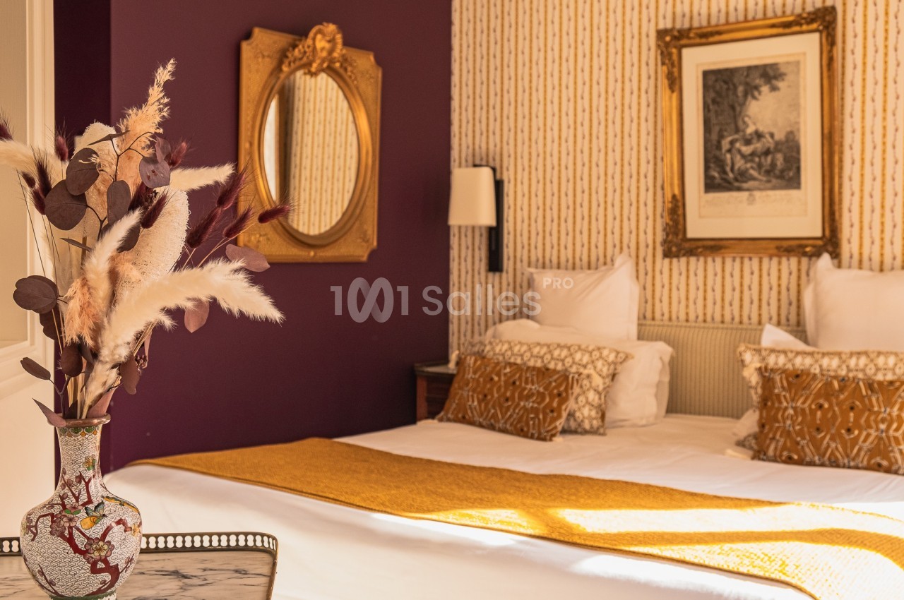 Chambre décorée avec des tons chauds, lit double, coussins dorés, miroir ovale et vase de fleurs séchées.