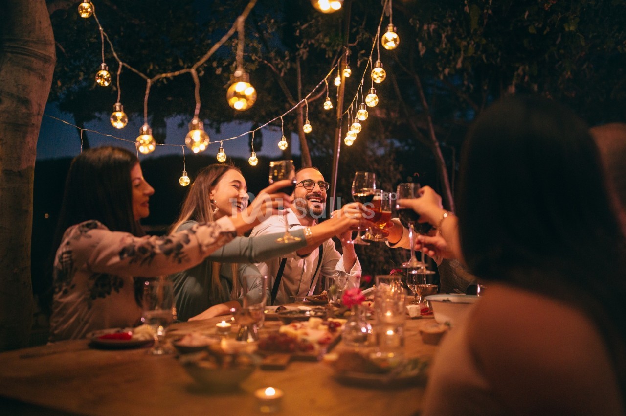 Un groupe de personnes trinque autour d'une table en extérieur, décorée de lumières suspendues, en soirée.