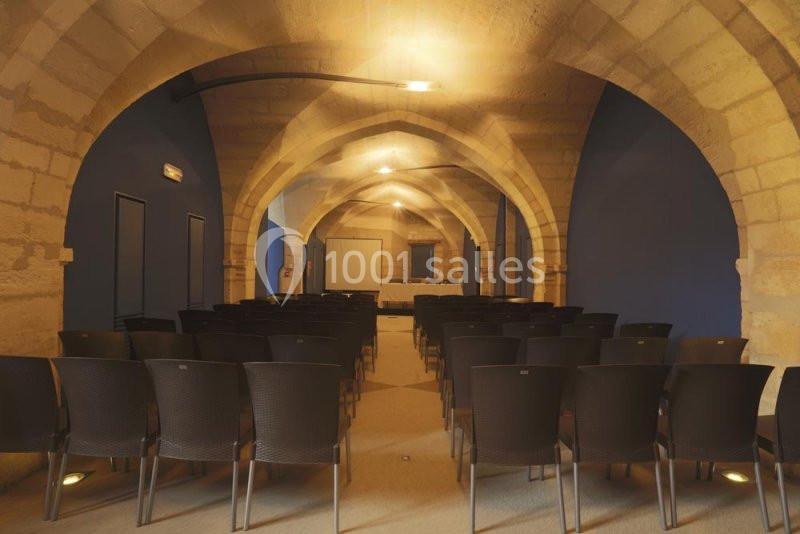 Salle voûtée en pierre avec rangées de chaises noires disposées face à un écran de présentation.
