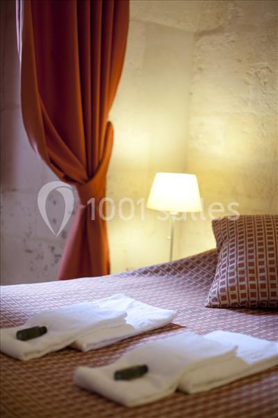 Lit avec couvre-lit à motifs, deux serviettes pliées avec savons, rideaux rouges et lampe allumée en arrière-plan.