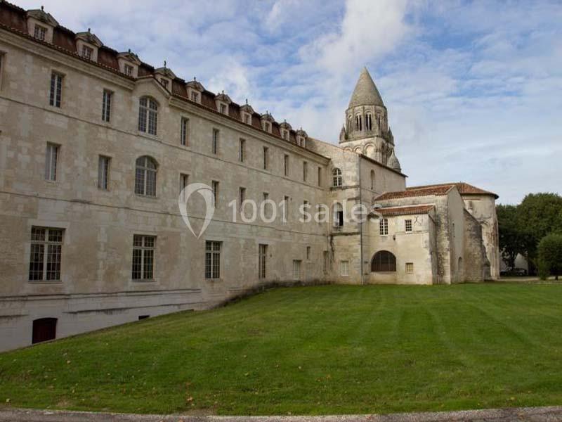 Bâtiment historique en pierre avec une tour conique, entouré d'une pelouse et d'arbres sous un ciel partiellement nuageux.