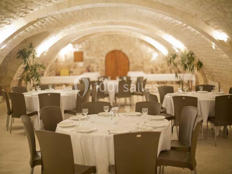 Salle voûtée en pierre avec tables rondes dressées pour un événement, éclairée par des lumières murales.