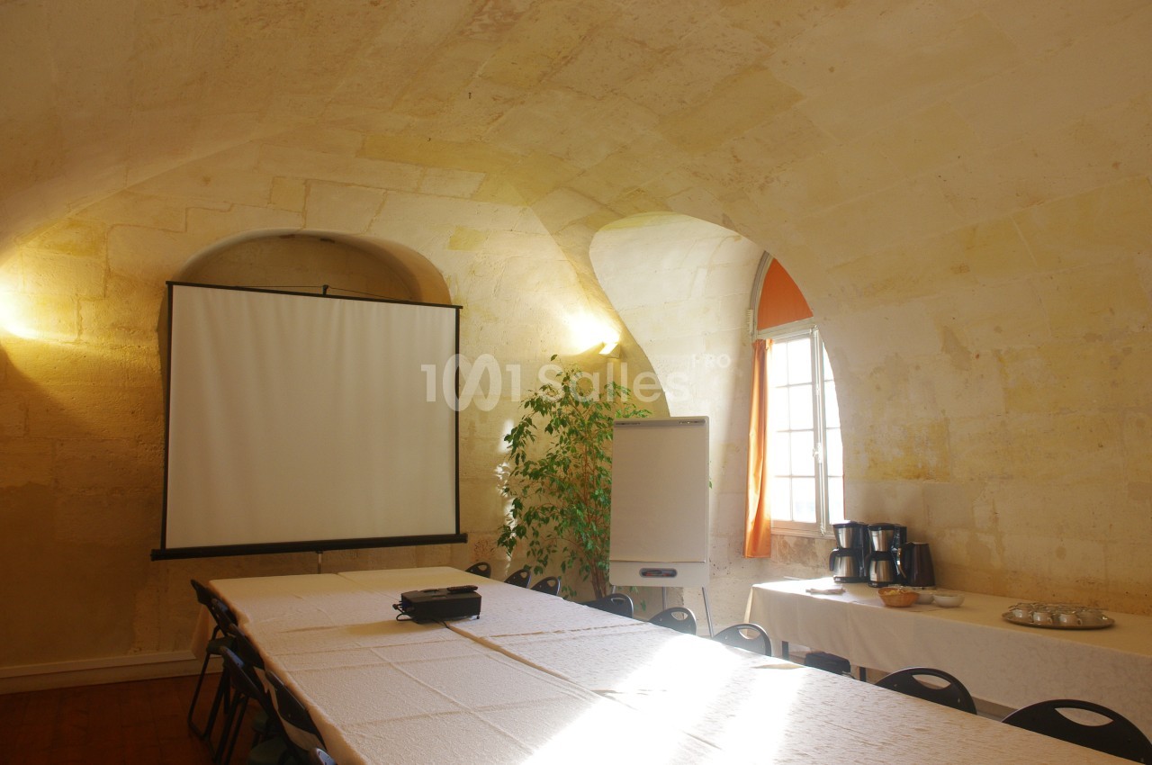 Salle voûtée éclairée avec écran de projection, tableau blanc, tables dressées et coin café sur le côté.