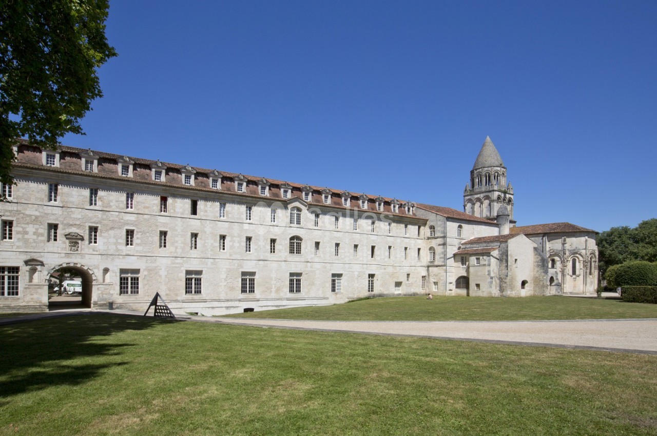 Façade d'un bâtiment historique en pierre avec une tour, entouré d'une pelouse et d'arbres sous un ciel bleu.
