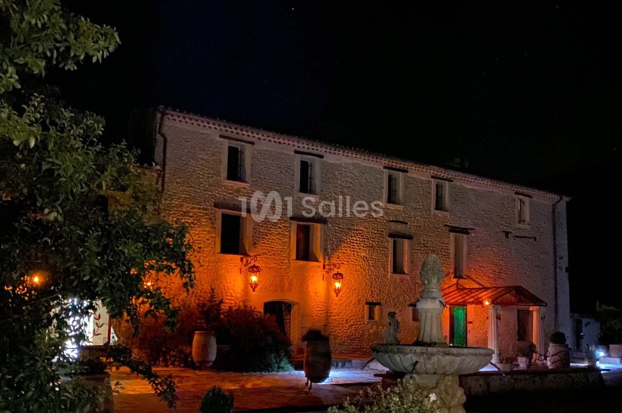 Façade d'une bâtisse en pierre éclairée de nuit, avec une fontaine au premier plan et des lanternes allumées.