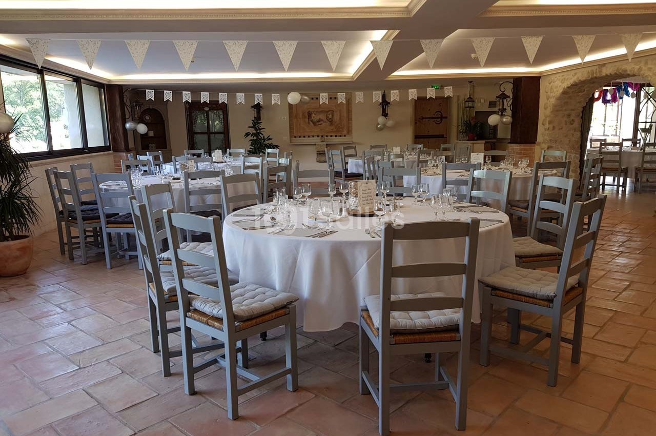 Salle de réception lumineuse avec tables rondes dressées, chaises en bois gris et décorations suspendues.