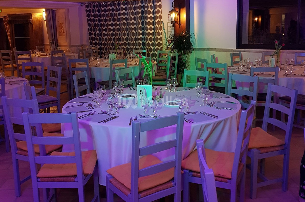 Salle de réception avec tables rondes dressées, nappes blanches, chaises en bois et éclairage coloré.