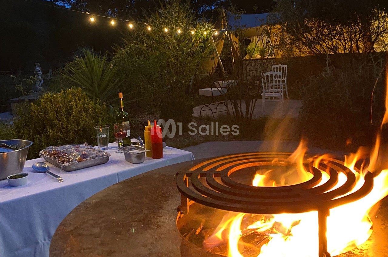 Barbecue allumé au premier plan avec une table dressée pour un repas en extérieur, éclairée par des guirlandes lumineuses.