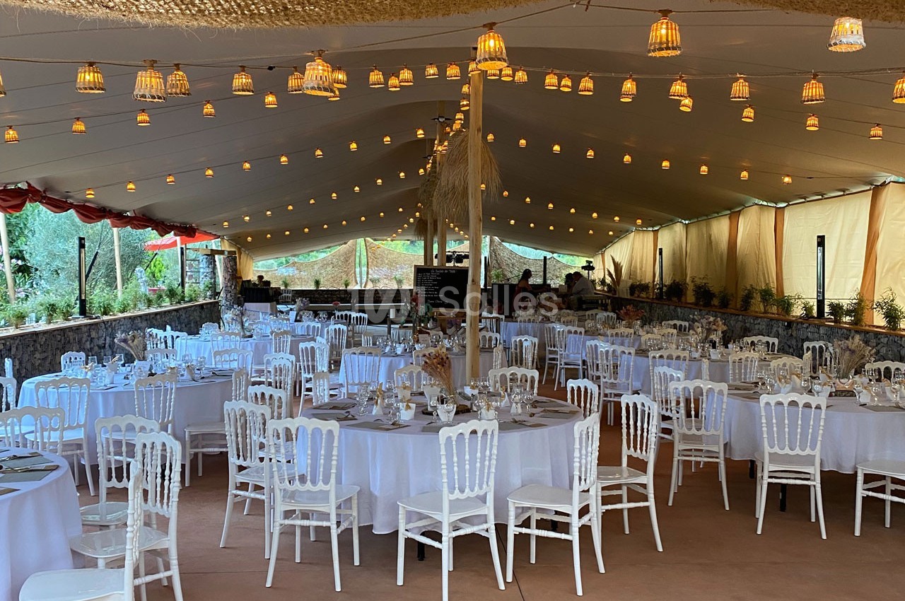 Salle de réception décorée avec des tables rondes dressées, des chaises blanches et des guirlandes lumineuses suspendues.