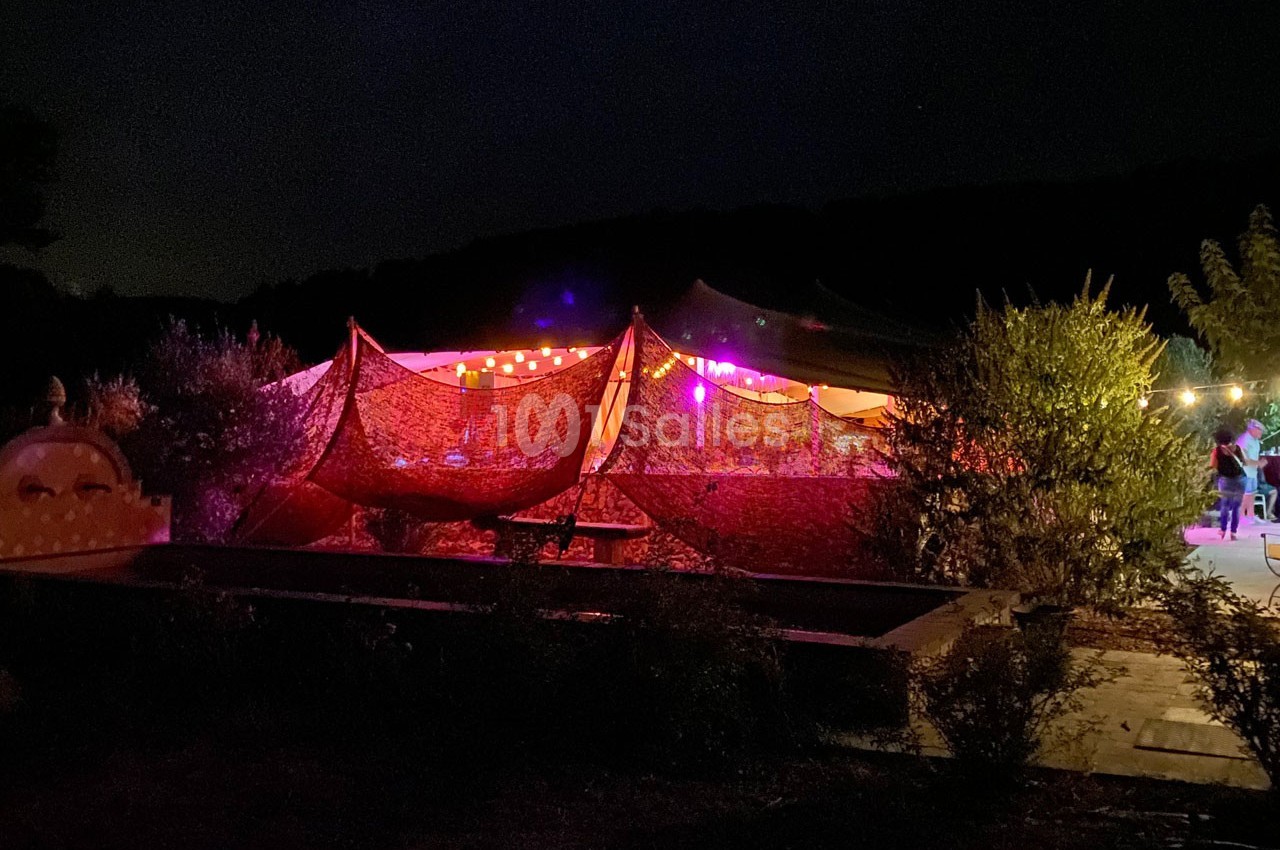 Tente éclairée par des guirlandes lumineuses dans un jardin sombre, entourée de végétation.