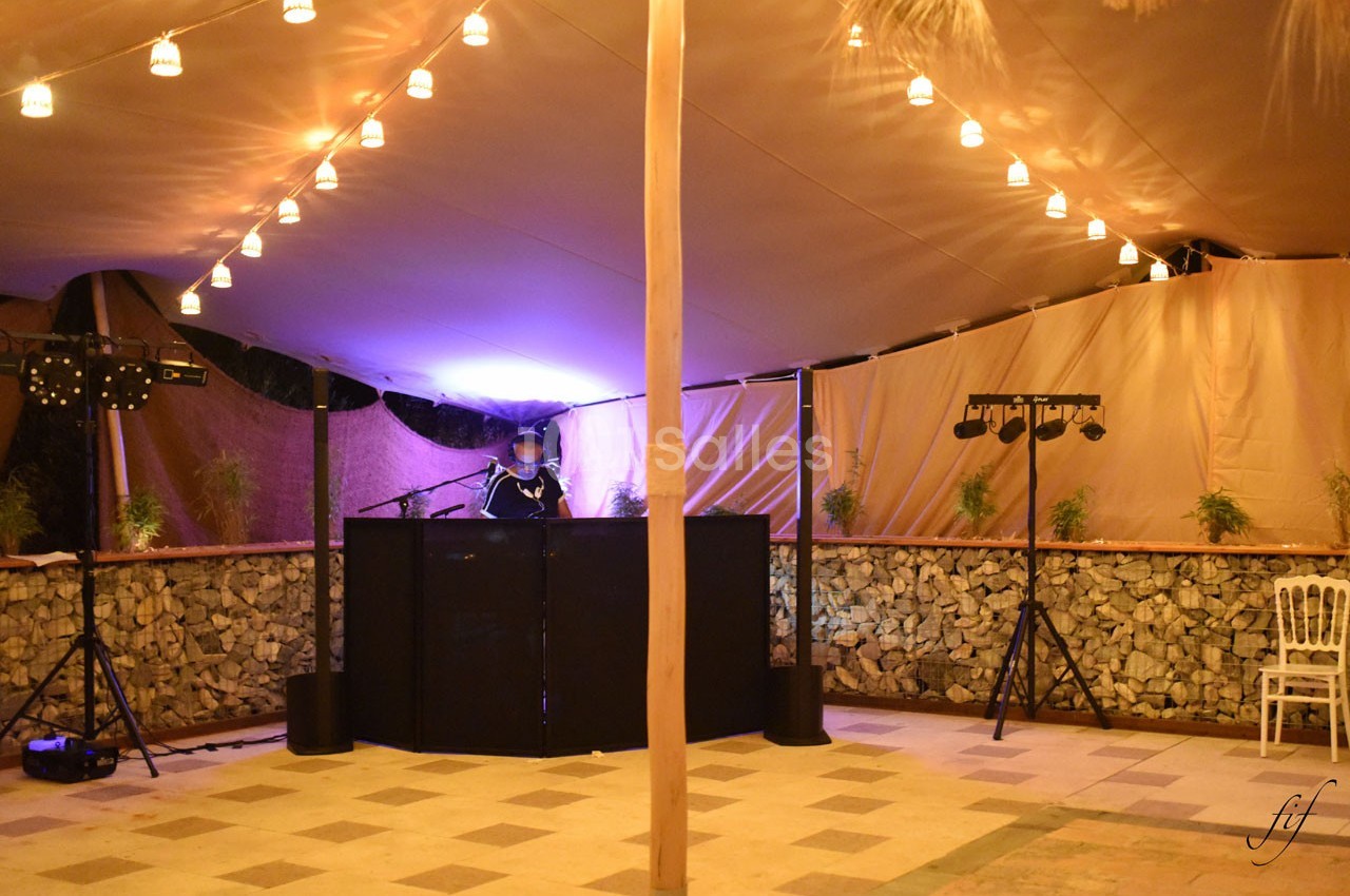 Un DJ mixe sous une tente éclairée par des guirlandes lumineuses, avec des équipements audio et une chaise vide à proximité.