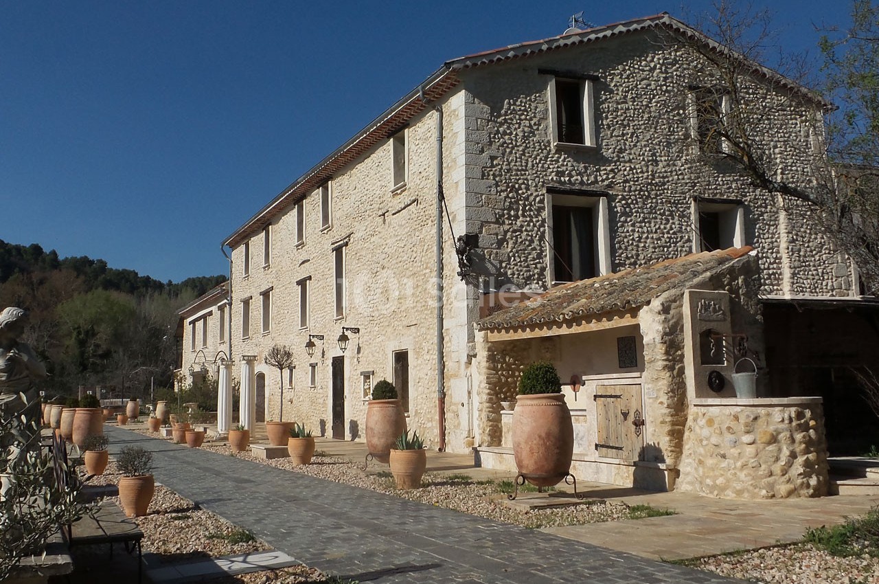 Bâtisse en pierre de style provençal avec chemin pavé, pots en terre cuite et végétation environnante.