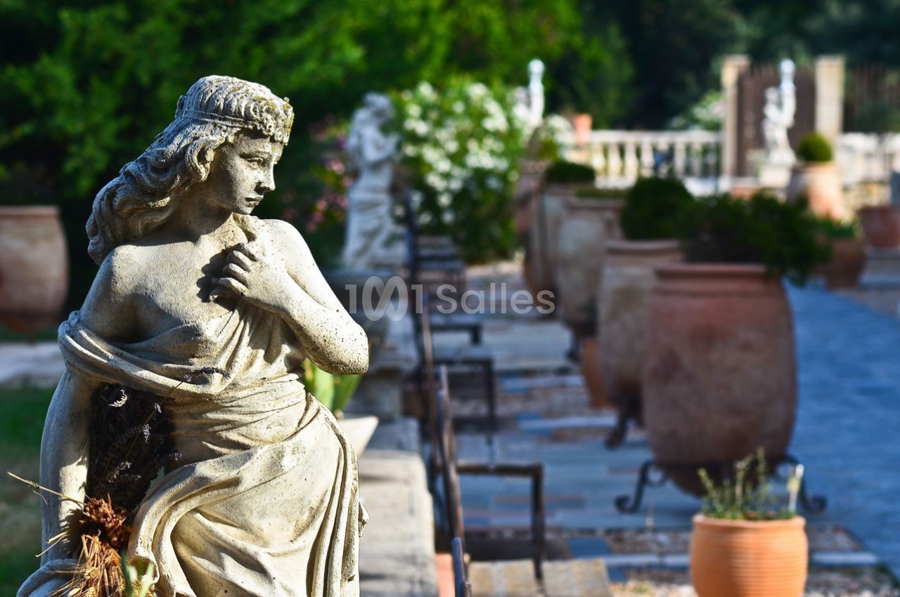Statue en pierre représentant une femme dans un jardin avec des pots en terre cuite et une allée pavée en arrière-plan.