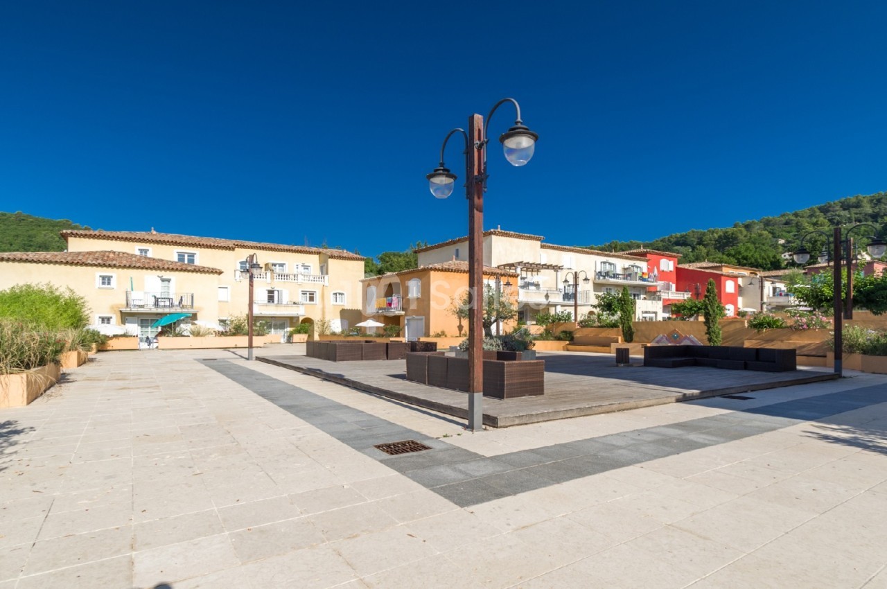 Place pavée bordée de bâtiments colorés et végétation, avec lampadaires et ciel bleu dégagé en arrière-plan.