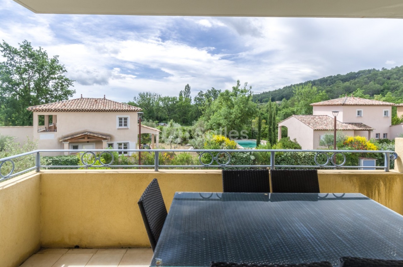 Vue depuis une terrasse avec table en verre et chaises, donnant sur des maisons, un jardin et des collines verdoyantes.