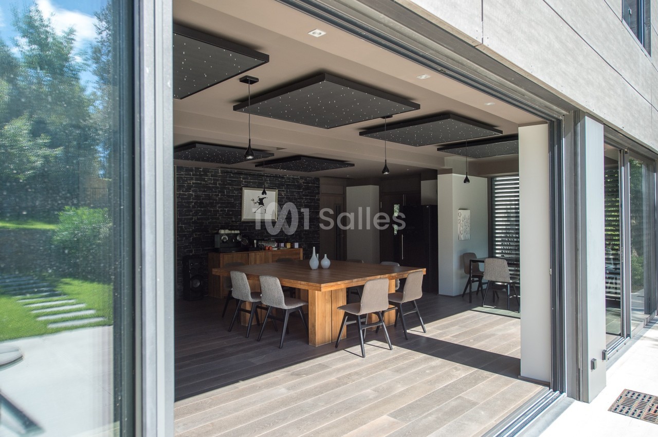 Salle à manger moderne avec une grande table en bois, chaises grises et baies vitrées donnant sur un jardin verdoyant.