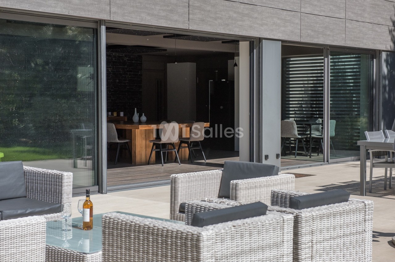Espace extérieur avec fauteuils en rotin et table basse, donnant sur une salle à manger moderne avec grandes baies vitrées.