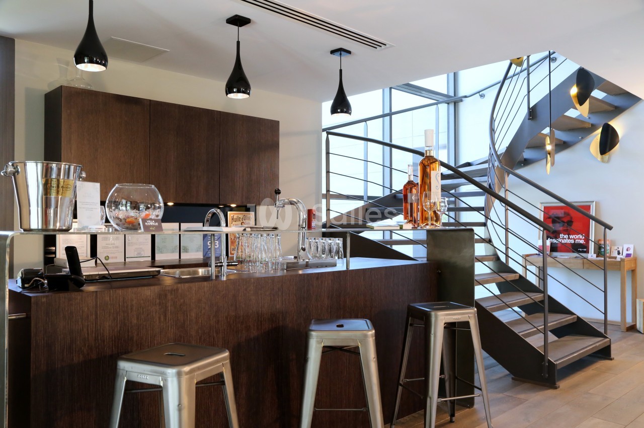 Bar moderne avec comptoir en bois sombre, tabourets métalliques, étagères avec bouteilles et escalier en arrière-plan.