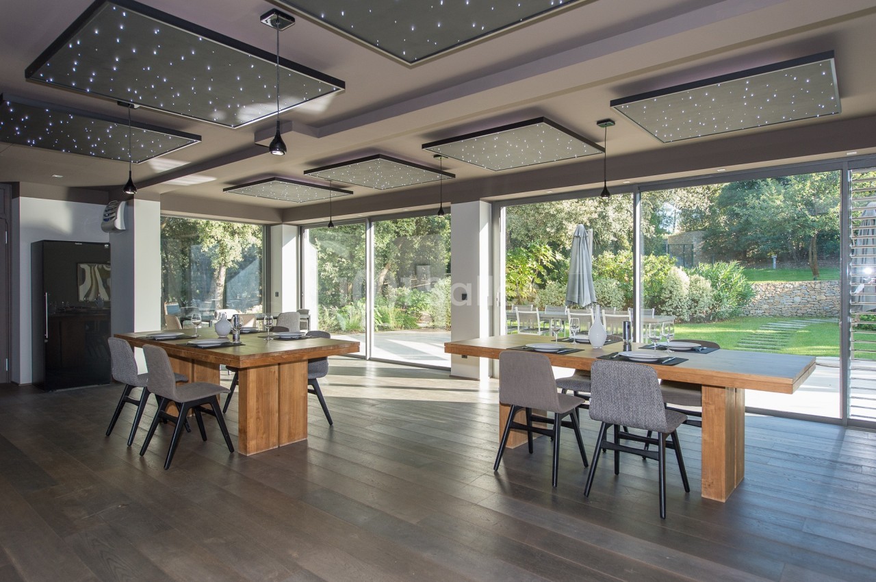 Salle à manger lumineuse avec grandes baies vitrées, tables en bois, chaises grises et vue sur un jardin verdoyant.