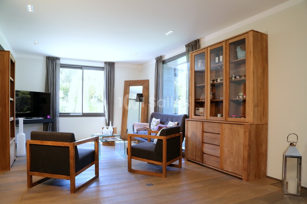 Salon lumineux avec mobilier en bois, fauteuils noirs, grande fenêtre et étagères vitrées contenant des objets décoratifs.