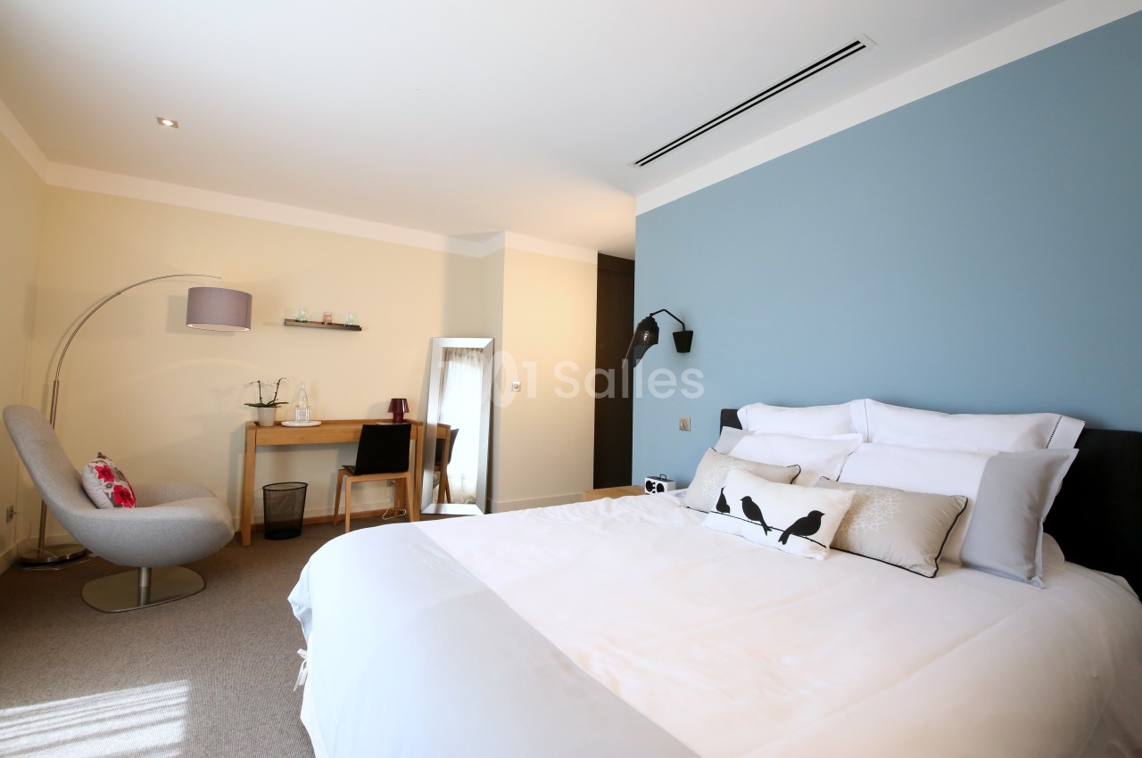 Chambre lumineuse avec lit double, mur bleu, bureau en bois, fauteuil gris et miroir sur pied.