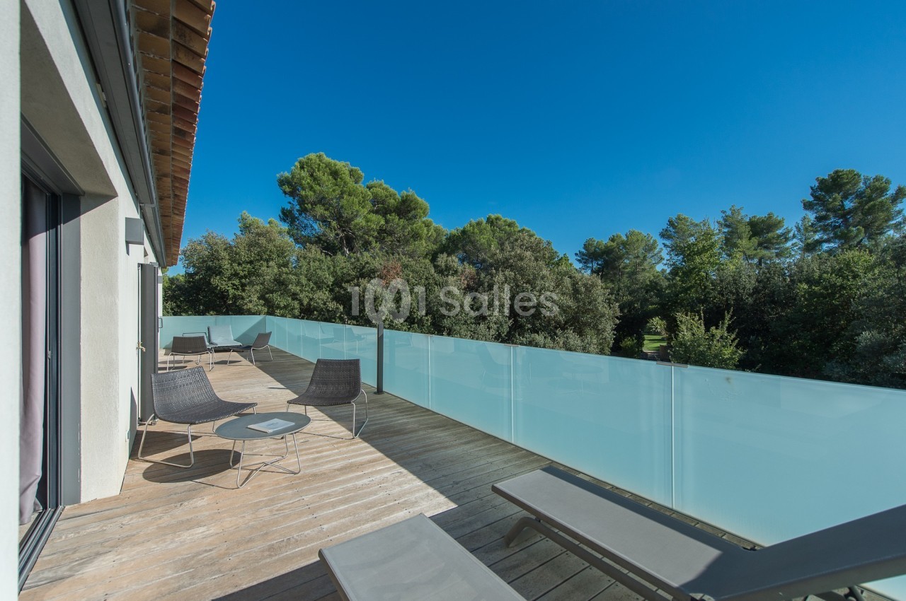 Terrasse en bois avec chaises longues et table, bordée d’un garde-corps en verre dépoli, entourée de végétation.