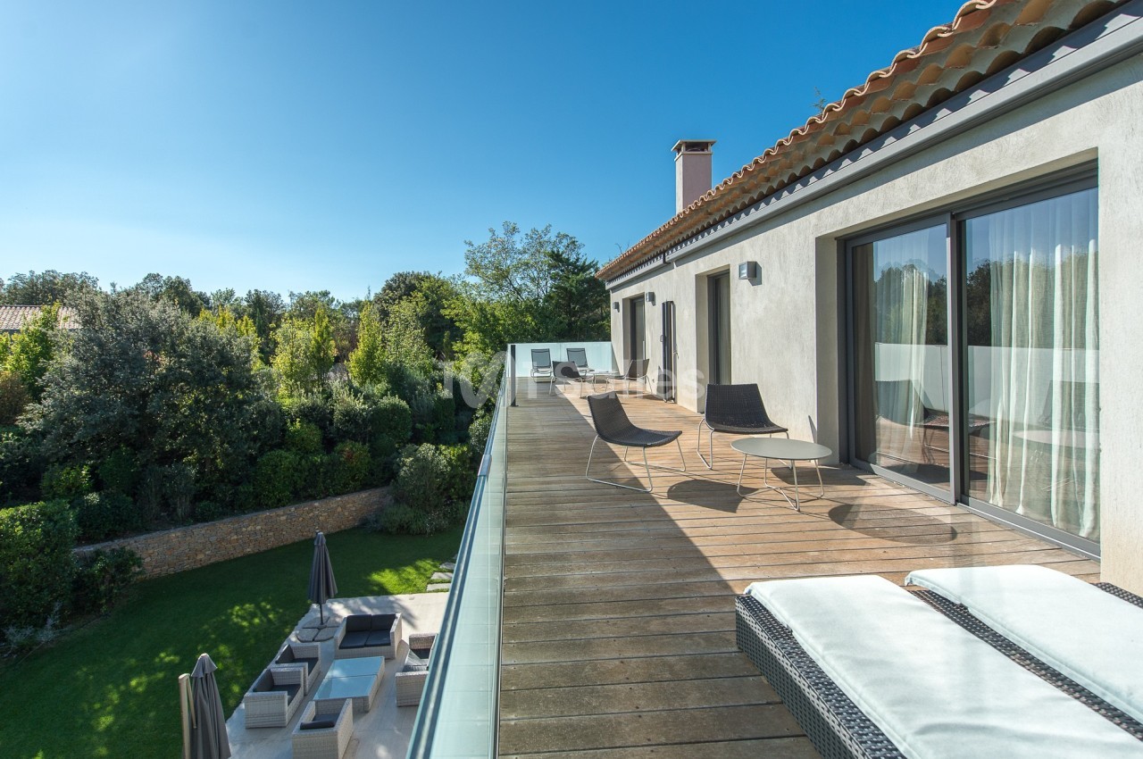 Terrasse en bois avec chaises longues et vue sur un jardin arboré, attenante à une maison moderne sous ciel dégagé.